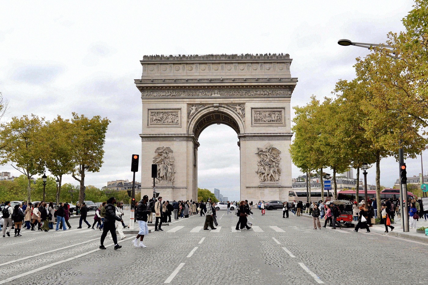 Des piétons traversent la rue devant l'Arc de Triomphe, bordé d'arbres aux couleurs automnales.