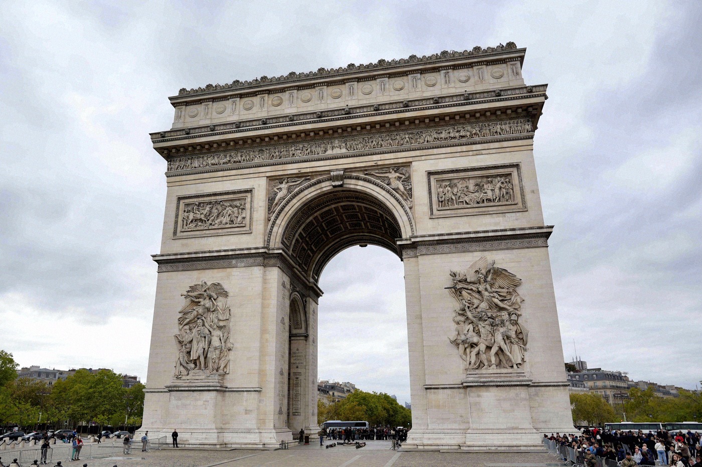 Gros plan sur l'Arc de Triomphe et ses reliefs détaillés sur les piliers, sous un ciel nuageux.
