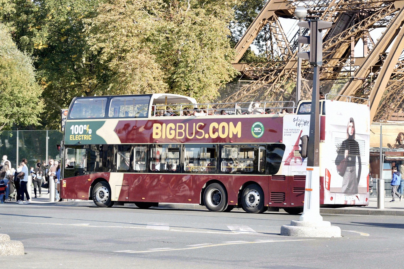 Un bus touristique rouge à impériale Big Bus s'arrête juste à côté de la structure en acier de la tour Eiffel, sur une avenue parisienne.