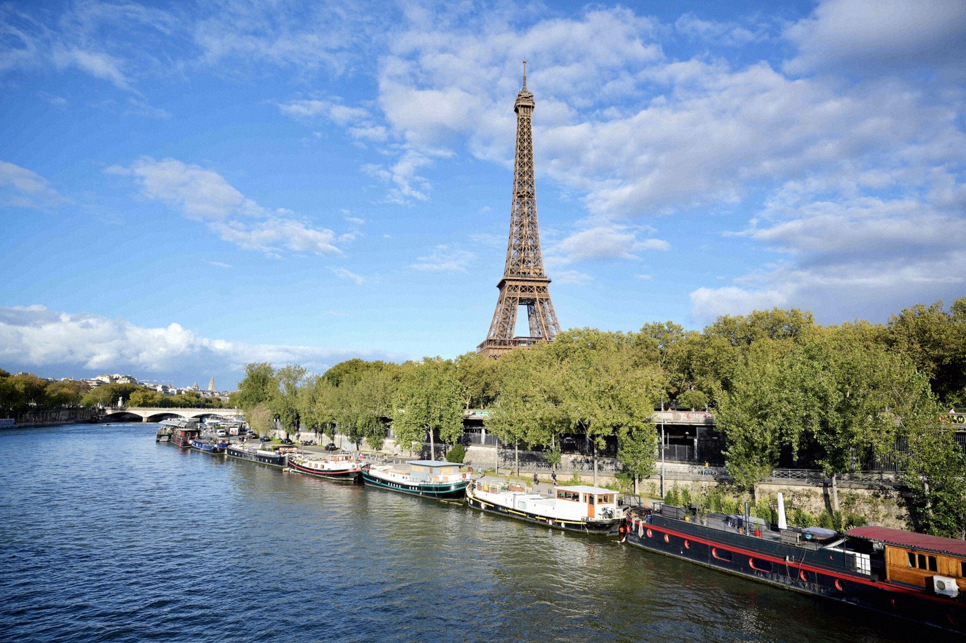 Vue depuis la Seine sur la tour Eiffel, avec des péniches alignées le long des berges végétalisées au premier plan.