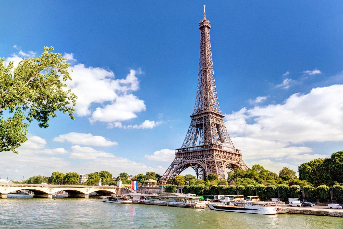 The Eiffel Tower photographed from the Seine in summer, with tour boats on the water and a bridge